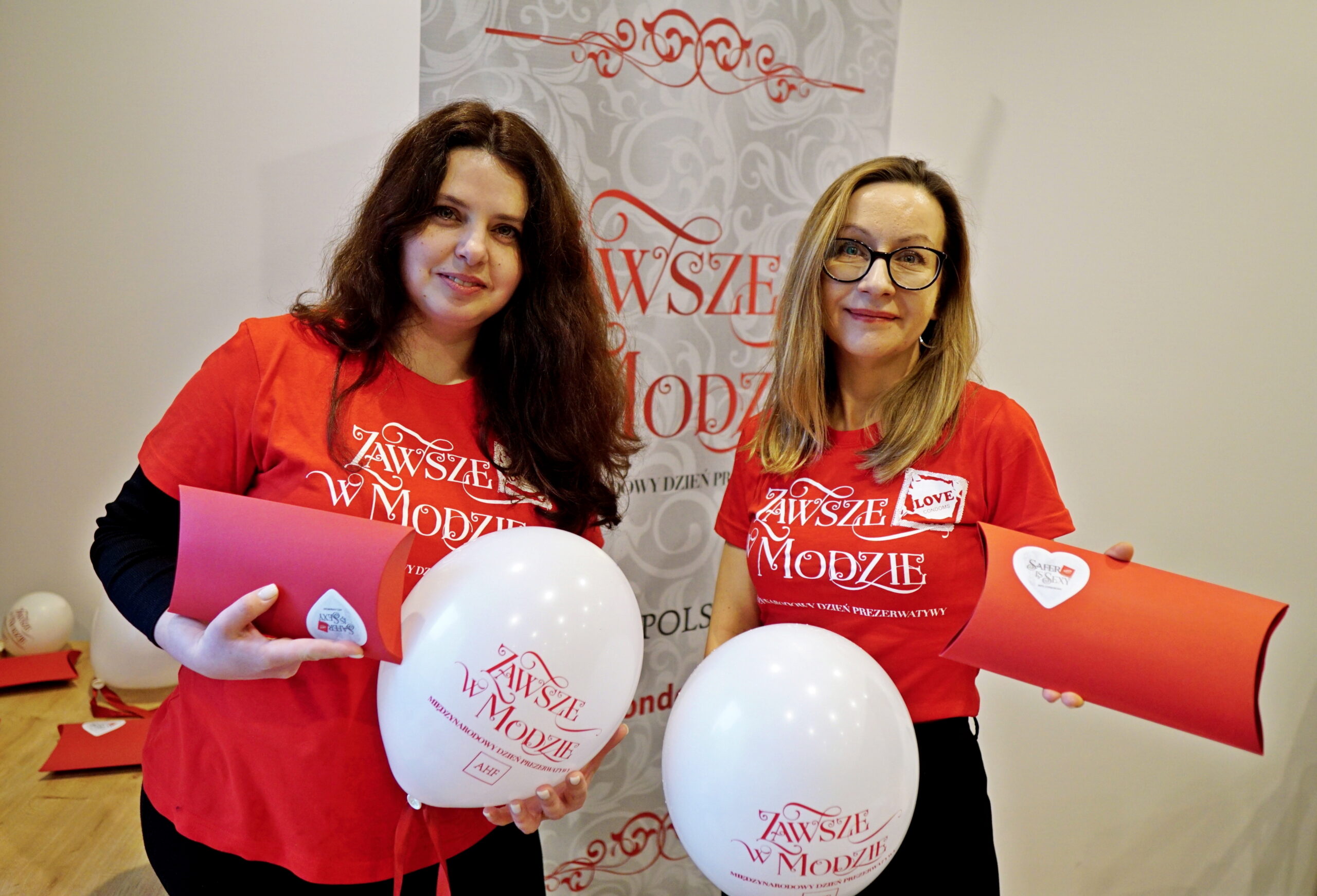 Two AHF Poland staff participating in a condom awareness campaign, holding educational materials and branded balloons during a sexual health promotion event.