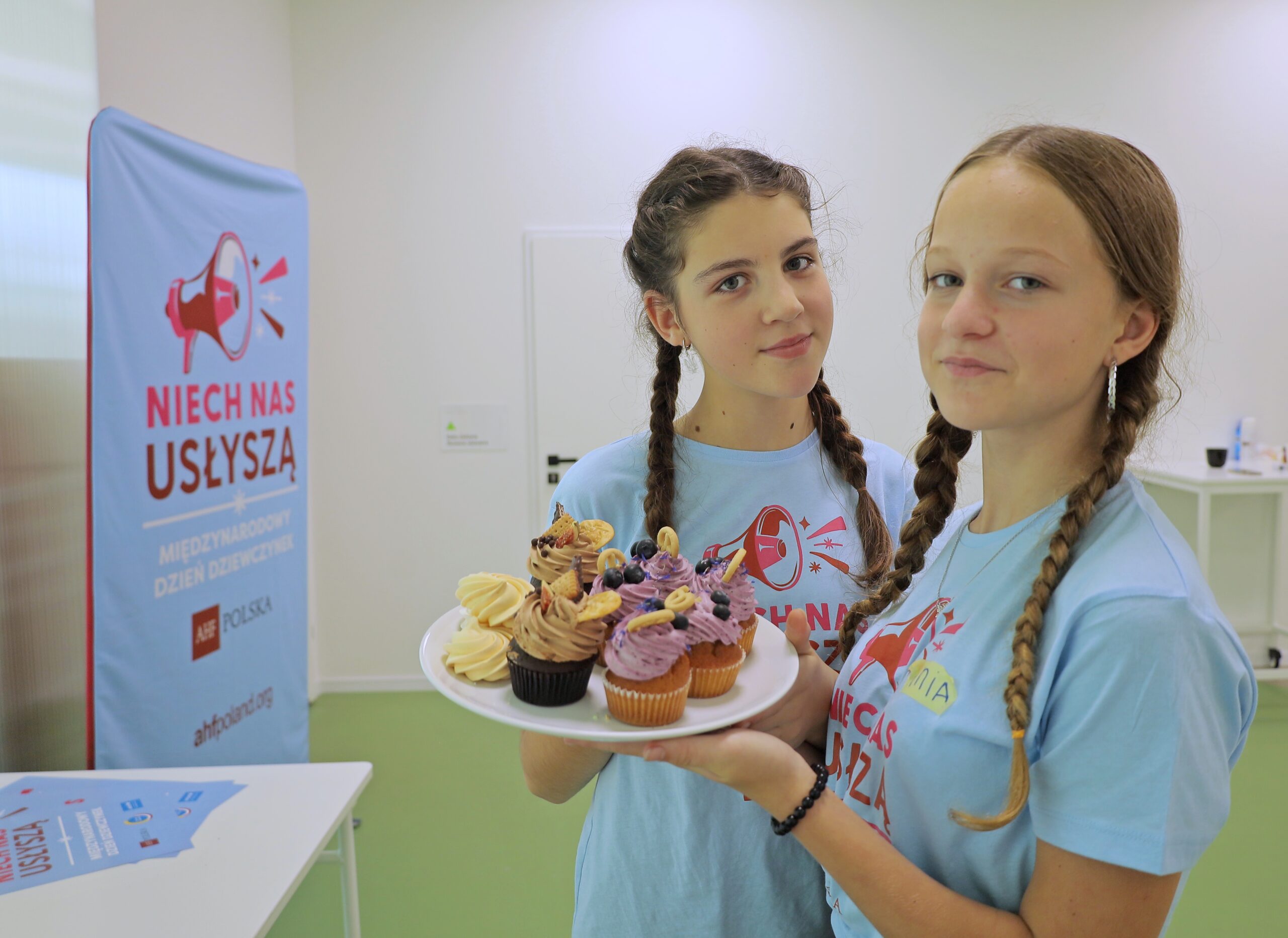 Two young participants holding a tray of cupcakes during an International Day of a Girl community event organized by AHF Poland.